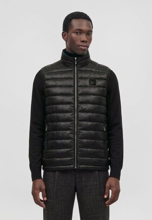 Un homme porte un gilet noir matelassé zippé sur un haut noir à manches longues, devant un fond clair et uni.