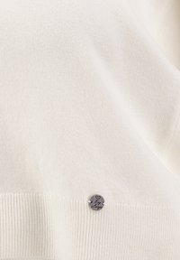 Maglione lavorato a maglia color crema con orlo a coste; presenta una piccola targhetta d'argento con testo inciso. Tessuto morbido con finitura liscia.