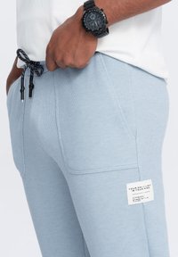 Vaaleansiniset verkkarit, jotka on valmistettu teksturoidusta kankaasta. Niissä on kiristysnyöri vyötäröllä, jossa on kontrastina mustat yksityiskohdat sekä brändilogo sivussa.