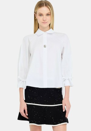 Witte blouse met knopen en een kraag, met driekwartmouwen en een metallic accenten. Gecombineerd met een zwarte, getextureerde rok met witte bies.