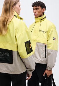 Δύο άτομα φορούν μπουφάν Tommy Jeans σε κίτρινο και μπεζ, το ένα κοιτά μπροστά κρατώντας μια μαύρη τσάντα, το άλλο δείχνει την πλάτη του.