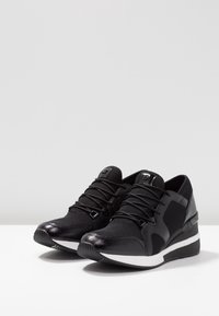 Chaussures de sport noires avec une tige en mesh, un design élégant, des accents en vernis texturé et une semelle intermédiaire blanche avec un talon compensé.