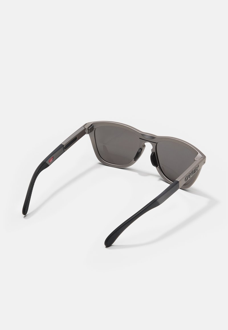 Gafas de sol con un marco de plástico gris y lentes de un gris oscuro. Presenta brazos de templo texturizados en negro con un acento de logo cerca de la bisagra.