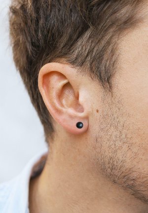 Schwarzes, rundes Steckerohrring in einem gestochenen Ohr, zusammen mit kurzem, strukturiertem braunem Haar und heller Hautfarbe präsentiert.