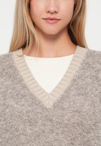 Lys grå, teksturert genser med V-hals og ribbet beige detaljer. Laget over en kremhvit, ribbet langermet topp.
