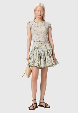 Ung kvinde med glat blondt hår iført kort blomstret top og plisseret blomstret nederdel, holder en beige clutch og har sorte sandaler på.