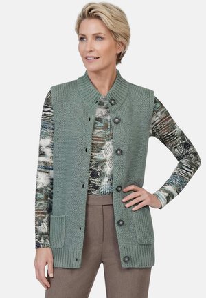 Vrouw draagt een groene gebreide mouwloze knoopvest over een langarm top met patroon en bruine broek, staand met één hand op de heup.