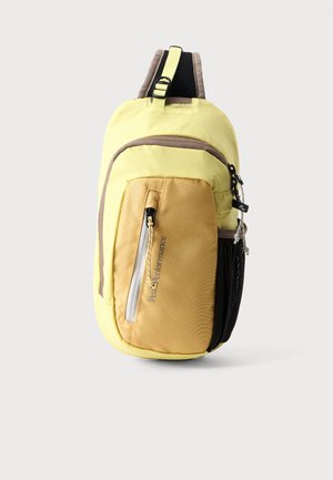 Kompakter gelber und beiger Sling-Rucksack mit einer vorderen Reißverschlusstasche und verstellbarem Schultergurt vor einfarbigem Hintergrund.