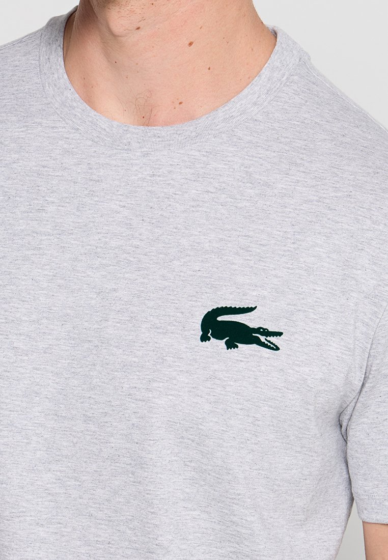 Homme portant un t-shirt gris clair avec un petit logo de crocodile vert foncé sur la poitrine gauche.