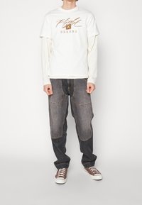 Kerman t-paita, jossa on "Flight"-teksti, valkoisen pitkähihaisen päälle puettuna, tummanharmaiden kuluneiden farkkujen ja ruskeiden lenkkarien kanssa.