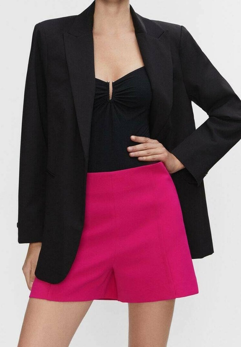 Femme portant un blazer noir avec un haut froncé noir et un short taille haute rose vif, debout avec une main sur la hanche.