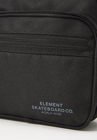 Zaino in tessuto nero con texture diagonale, dotato di tasca con zip e testo del logo blu "ELEMENT SKATEBOARD CO. WORLD WIDE" sulla parte anteriore.