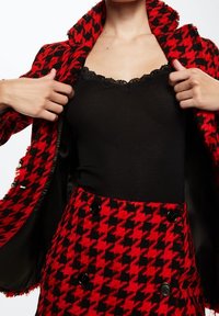 Rood en zwart houndstooth blazer met rafelige randen, gecombineerd met een zwart ribbel tanktop met kantafwerking, en een bijpassende rok met knopen.