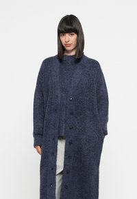 Lang, marineblå cardigan laget av mykt teksturert stoff. Den har knappelukking foran, vide ermer og sidelommer. Brukes over en grå topp.