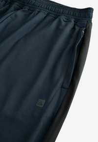 Pantalones deportivos de color azul marino con cintura elástica, bolsillo con cremallera, paneles laterales y un pequeño parche con logo. Textura suave.