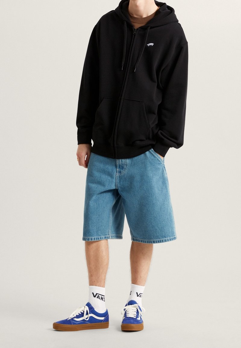 Personne portant un sweat à capuche noir zippé, un short en jean bleu clair, des chaussettes blanches Vans et des baskets Vans bleues avec des semelles marron.