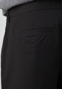 Zbliżenie spodni garniturowych w kolorze czarnym z jedną kieszenią z wypustką z tyłu oraz szarym swetrem częściowo widocznym w okolicy talii.
