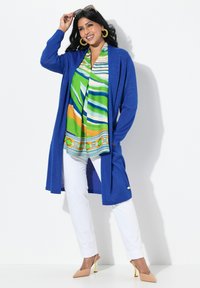 Langsleeve blauwe cardigan met subtiele sprankeling, gecombineerd met een groen-witte gebloemde blouse en witte broeken. Het model draagt beige hakken.