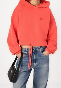 Sudadera corta color coral con dobladillo ajustable con cordón y puños elásticos, combinada con jeans azul claro. Un pequeño bolso negro añade contraste.