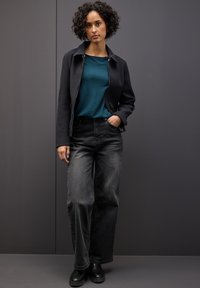 Schwarze, mit Reißverschluss versehene Jacke, tealfarbener, strukturierter Pullover, schwarze weit geschnittene Jeans und schwarze Stiefel. Model steht vor einem grauen Hintergrund.