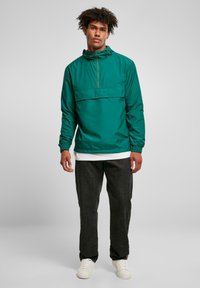 Grøn letvægts pullover-jakke med en kvartlang lynlås, frontlomme og fast hætte. Bæres over en hvid skjorte og kombineres med mørke jeans.
