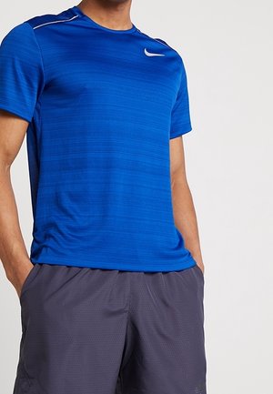 Mand iført en blå Nike kortærmet atletiktrøje og mørkegrå shorts med hænderne i lommerne, vist fra nakke til midt på låret.