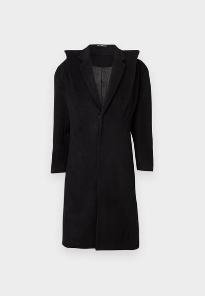 Palton lung negru cu umeri structurați lățit, guler tip rever și închidere frontală, agățat pe un fundal alb.