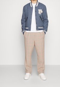 Blå ullblandning varsityjacka med vita ränder och broderad logotyp, kombinerad med beige rutiga byxor och vita sneakers.