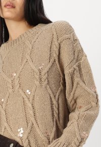 Pulover bej tricotat cu un model de tricotaj cu cabluri, accentuat cu paiete pe mâneci, design cu guler rotund și detalii texturate.