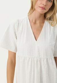 Vit klänning med V-ringning, korta puffärmar, texturerat tyg och sammanknuten midja för ökad volym.