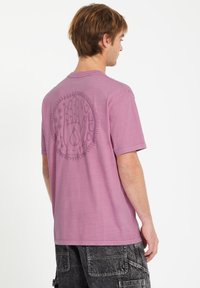 Lilla bomulds T-shirt med korte ærmer, featuring et håndgrafik og cirkulært design på ryggen, og en afslappet pasform.