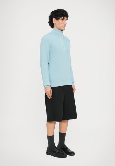 Pull à fermeture éclair bleu clair en matière texturée, associé à un short noir et des mocassins noirs, posé devant un fond blanc.