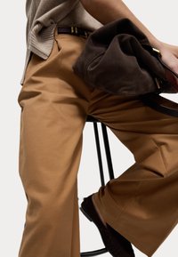 Bruna vida byxor gjorda av ett slätt tyg, i kombination med en mörkbrun mockaväska. Bältet är av mörkt läder med metalliska detaljer.