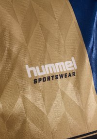 Guldt stof med et tekstureret diamantmønster; har mærket "hummel" i hvidt og "SPORTSWEAR" i sort. Blå accenter til stede.