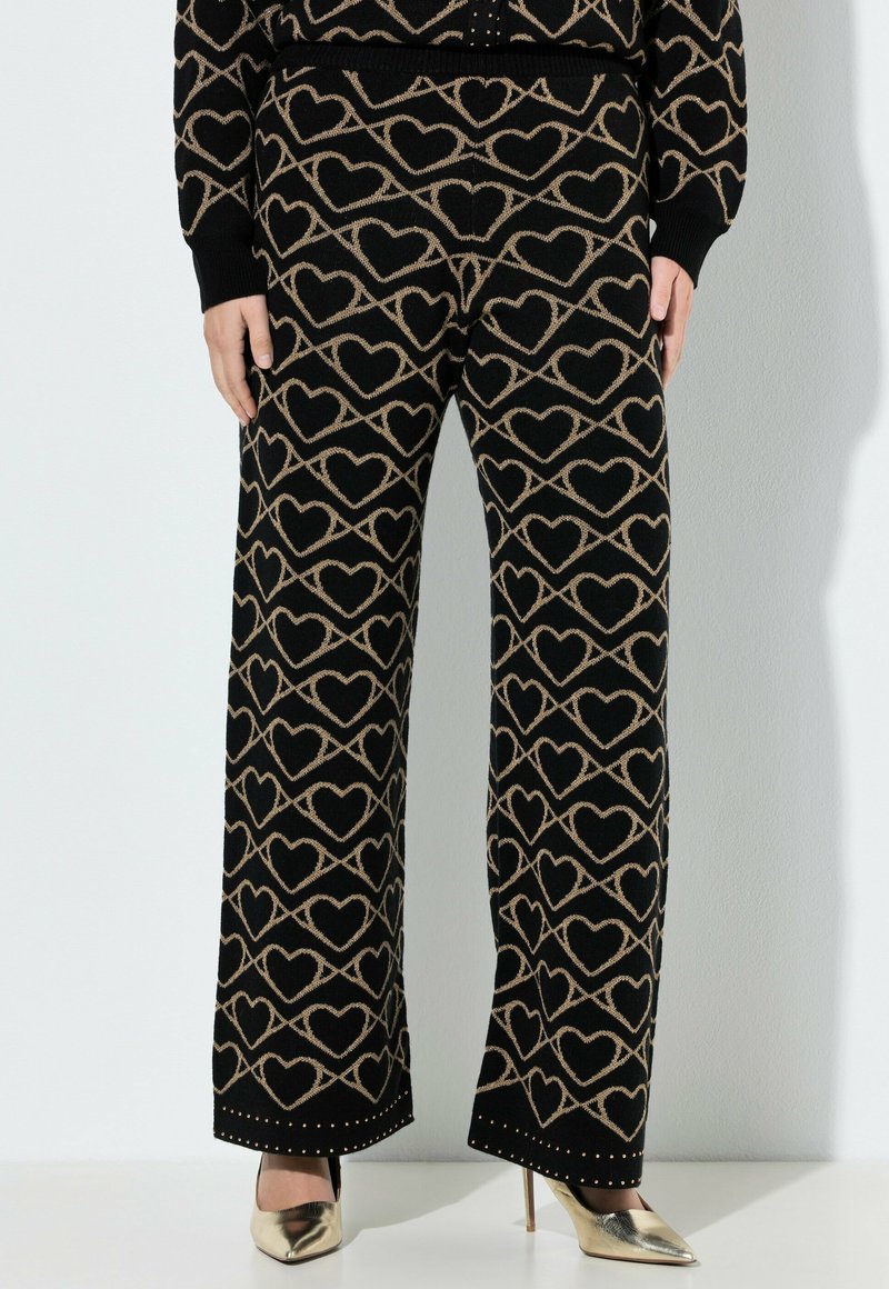 Sorte strikbukser med beige hjerte- og uendelighedsmønster, kombineret med guldsko med høje hæle og en matchende mønstret top.