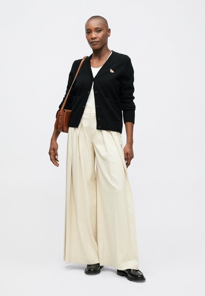 Cárdigan negro con escote en v y bolsillos frontales, combinado con pantalones plisados de talle alto en color marfil y zapatos negros. Se incluye un bolso de hombro marrón.