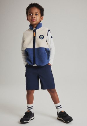 Jonge jongen staat met handen in zijn zakken, draagt een blauw-wit fleecevest, wit shirt met lange mouwen, marineblauwe korte broek, gestreepte sokken en zwarte sneakers.