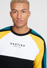 Sweater colorido com painéis pretos, brancos, amarelos e teal. Apresenta um decote redondo e punhos canelados, com o logótipo "KAOTIKO" na parte frontal.
