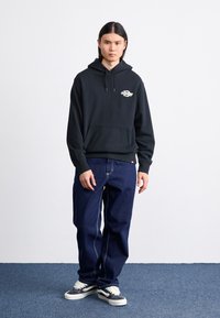Sort bomulds hoodie med en lomme foran og logodetalje, parret med blå denimjeans. Modellen har grå sneakers med hvide snørebånd på.