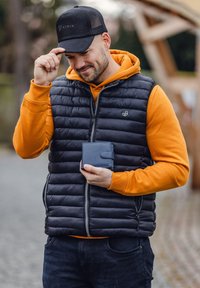 Black puffy vest over an orange hoodie, holding a blue leather wallet. Cap featuring "STRIX" logo. Outdoor setting with stone pavement.