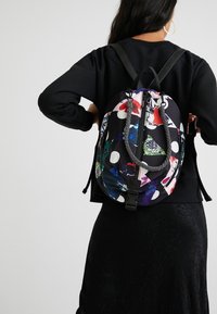 Sac à dos noir rond avec un imprimé abstrait coloré, incluant des pois et des formes géométriques, doté de bretelles noires et d'une fermeture supérieure.