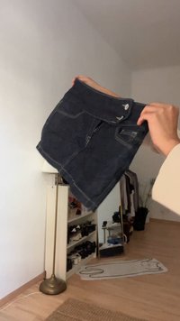Ruky držia tmavú džínovú sukňu s prednými vreckami a gombíkmi v miestnosti s podlahovou lampou, policami na topánky a kobercom v tvare mačky.