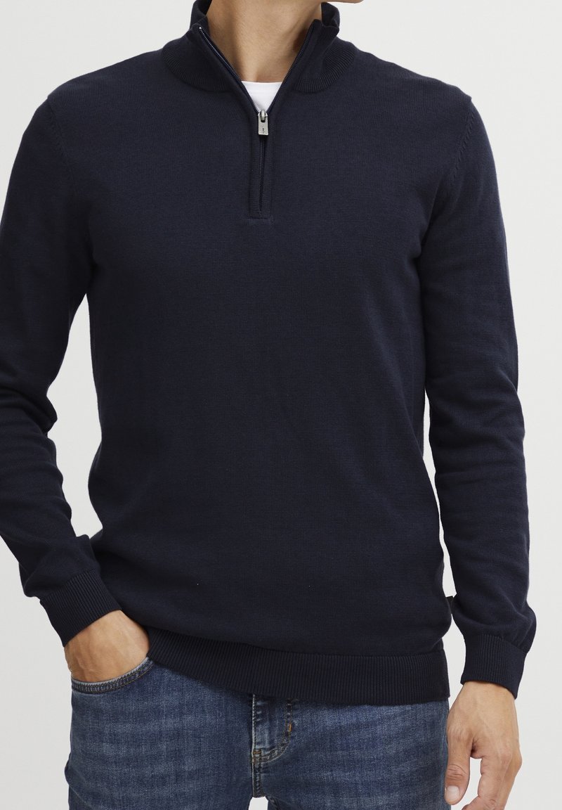 Homme portant un pull bleu marine foncé à fermeture éclair quart sur un t-shirt blanc, une main dans la poche d'un jean bleu, sur un fond uni.