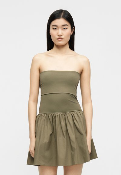 Γυναίκα με ίσια μαύρα μαλλιά, φορώντας αμάνικο ελιάς πράσινο φόρεμα με εφαρμοστό μπούστο και φούστα με πιέτες, στέκεται μπροστά σε απλό φόντο.