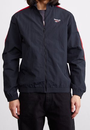 Osoba ubrana w czarną kurtkę Reebok zapinaną na zamek błyskawiczny, z czerwonymi paskami na rękawach i elastycznymi mankietami, w połączeniu z czarnymi spodniami.