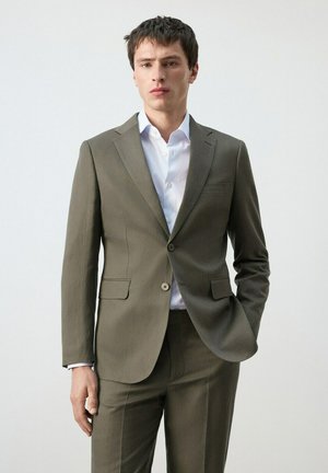 Uomo che indossa un abito verde oliva e una camicia bianca, con una mano in tasca, in piedi contro uno sfondo chiaro e uniforme.