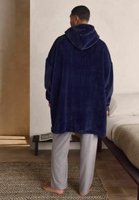 Tumesinine fliisist kapuutsiga sviiter, millel on suur kapuuts ja oversized sobivus, koos heledate hallide sirgete pükstega. Taust on neutraalne.