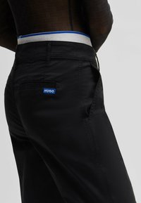 Pantalons noirs au tissu lisse, dotés d'une ceinture blanche avec des accents bleus et d'un petit logo HUGO bleu sur la poche arrière.