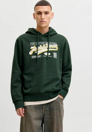 Młody mężczyzna w ciemnozielonej bluzie z kapturem Jack & Jones z tekstem i logo, z jedną ręką w kieszeni, stoi na jednolitym białym tle.