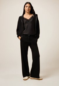 Sweat à fermeture éclair noir sur un débardeur en satin noir avec des bordures en dentelle, associé à un pantalon large noir et des baskets blanches.